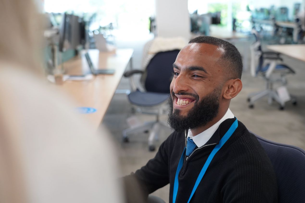 Smiling man with a beard, wearing a black sweater and blue lanyard, conversing with a colleague in a bright office.