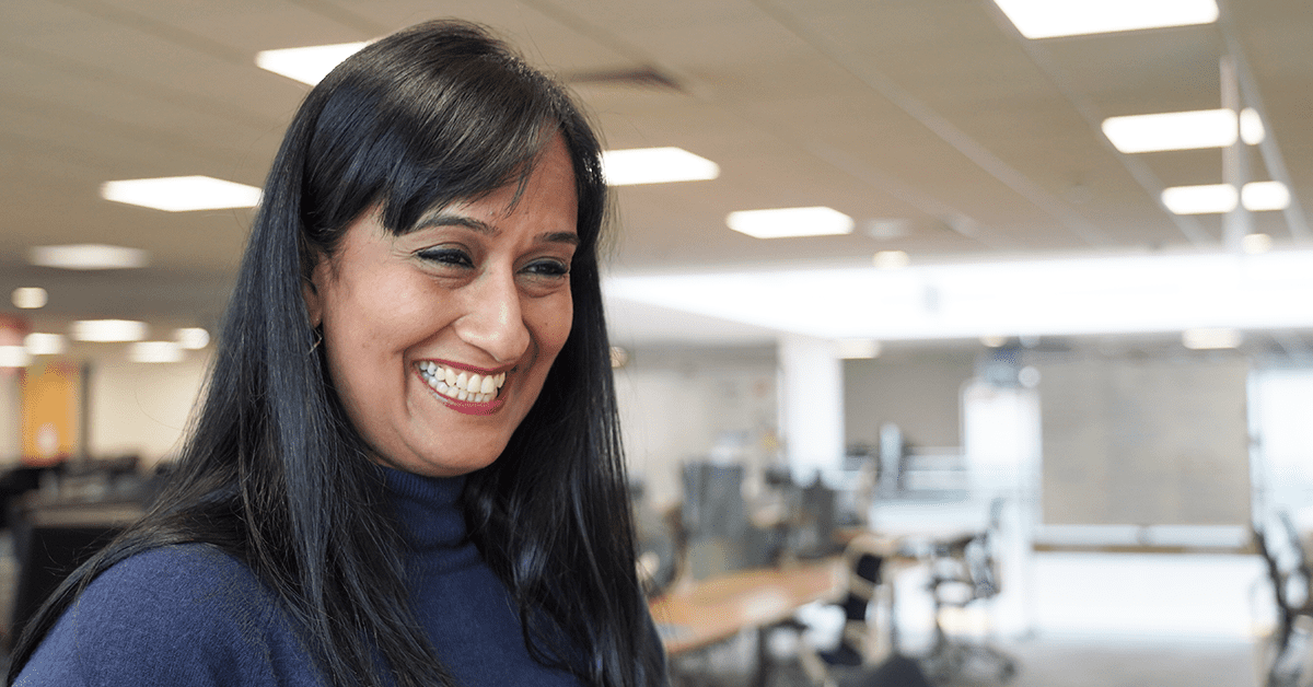 Smiling woman in a blue top with long black hair, standing in an office environment.