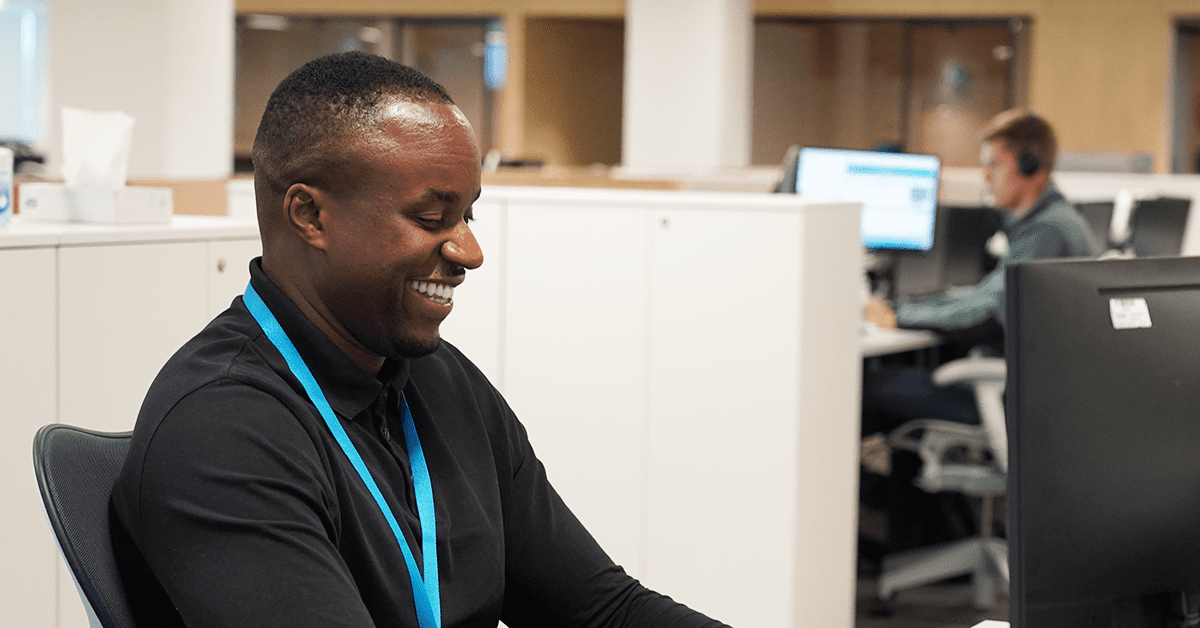 Joyful man in a black shirt and blue lanyard seated at an office desk.